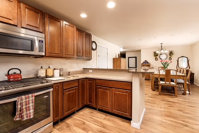 A spacious kitchen with wooden cabinets, a stainless steel microwave and oven, a red kettle on the stove, and a countertop with various containers. The kitchen opens into a dining area with a wooden dining table, chairs, a wall clock, and decorative wall hangings. The floor is wooden and the space is well-lit with ceiling lights.