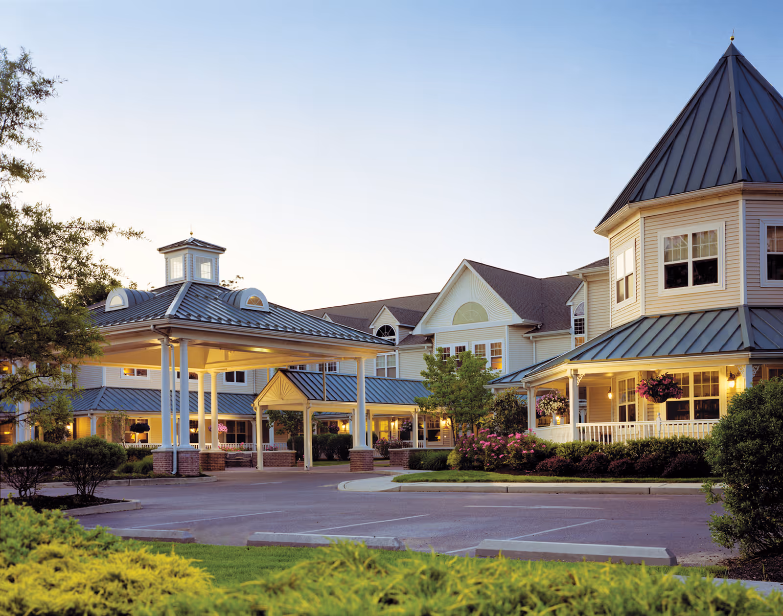 Exterior view of Brandywine Brandall Estates by Monarch, showing a large building with a covered entrance, multiple windows, and a landscaped garden with bushes and flowers in front. The sky is clear and it appears to be early evening.