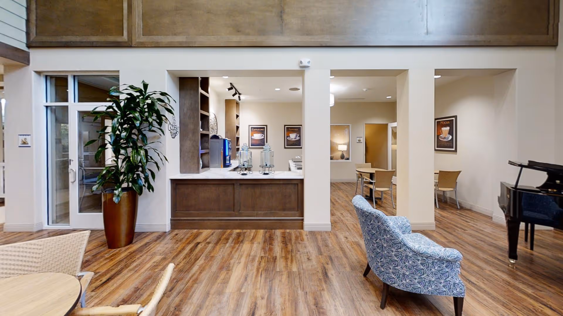 Interior view of a senior living facility common area with wooden flooring, a large potted plant, a blue patterned armchair, a black grand piano, and a counter area with water dispensers and coffee machines. There are tables and chairs in the background and framed coffee-themed artwork on the walls.