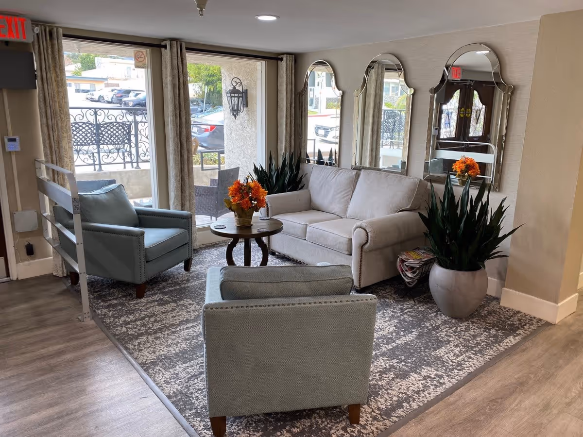 A cozy senior living facility lounge area with a beige sofa, two light blue armchairs, a round wooden coffee table with a flower arrangement, large potted plants, three decorative mirrors on the wall, and large windows showing an outdoor parking area.