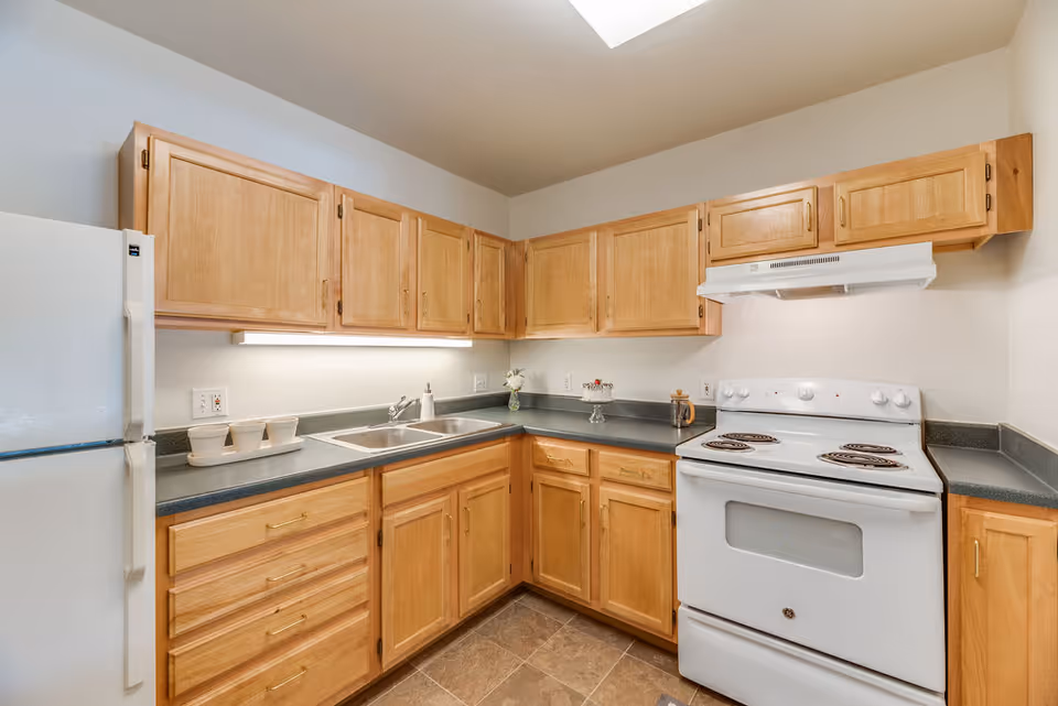 A clean kitchen with light wood cabinets, a white refrigerator, a white electric stove with an oven, a double sink, and gray countertops. The kitchen has tiled flooring and a skylight above providing natural light.