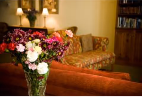 A cozy living room with a floral-patterned sofa, a glass vase filled with colorful flowers in the foreground, warm lighting from table lamps, and a bookshelf filled with books in the background.