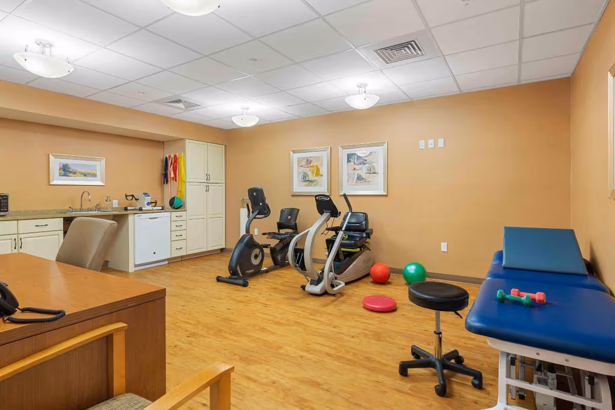 A therapy or exercise room with two stationary exercise bikes, colorful therapy balls, small dumbbells on a blue padded therapy table, a rolling stool, a wooden desk with a chair, and a kitchenette area with cabinets, a sink, and a mini fridge. The walls are beige with framed artwork, and the floor is wood.