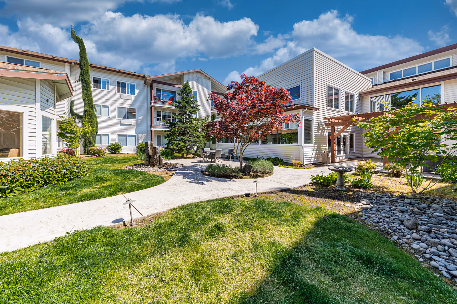 Outdoor courtyard area of a senior living facility with a paved walkway, green grass, various trees and shrubs, and a seating area with a table and chairs. The building surrounding the courtyard is white with multiple windows and balconies under a partly cloudy blue sky.