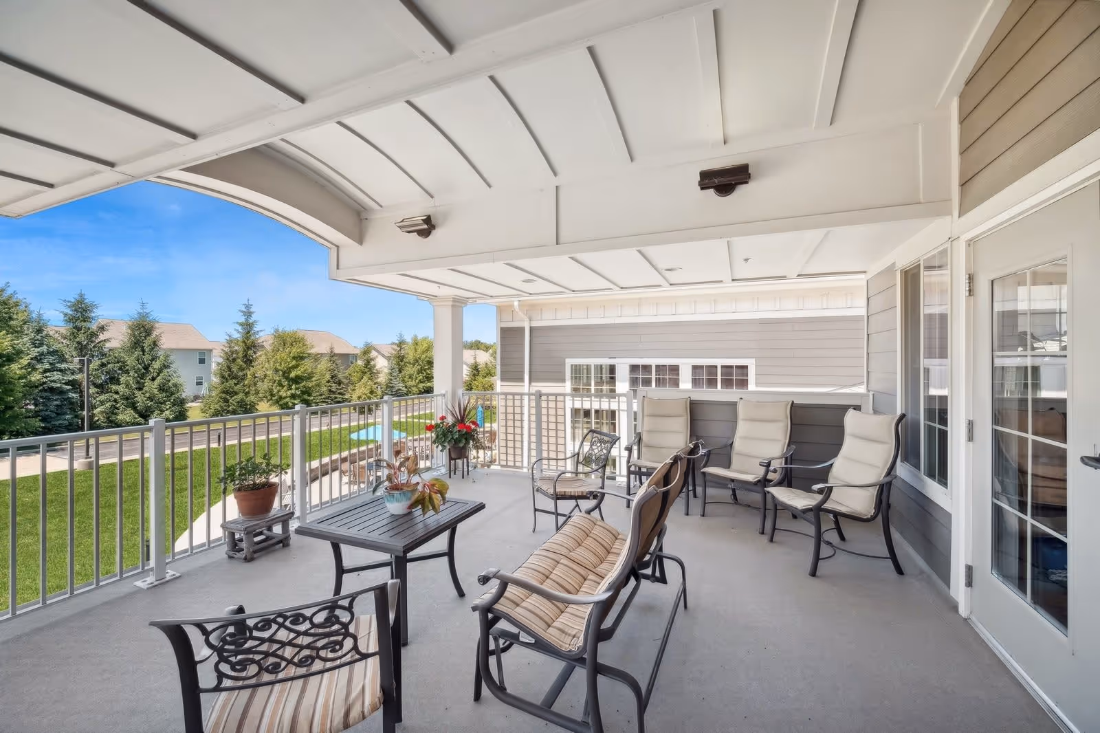 Covered outdoor patio area with cushioned chairs, a small table with potted plants, and a view of green grass, trees, and neighboring buildings under a clear blue sky.