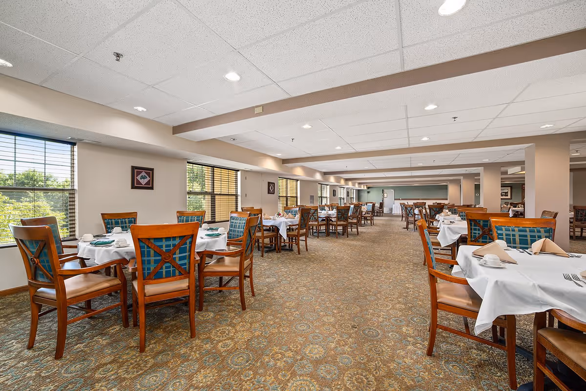 A spacious dining room with multiple tables covered in white tablecloths, each set with cups, plates, napkins, and silverware. Wooden chairs with blue and green patterned cushions surround the tables. Large windows on the left side allow natural light to fill the room, and the ceiling has recessed lighting. The carpet has a detailed pattern in earth tones.