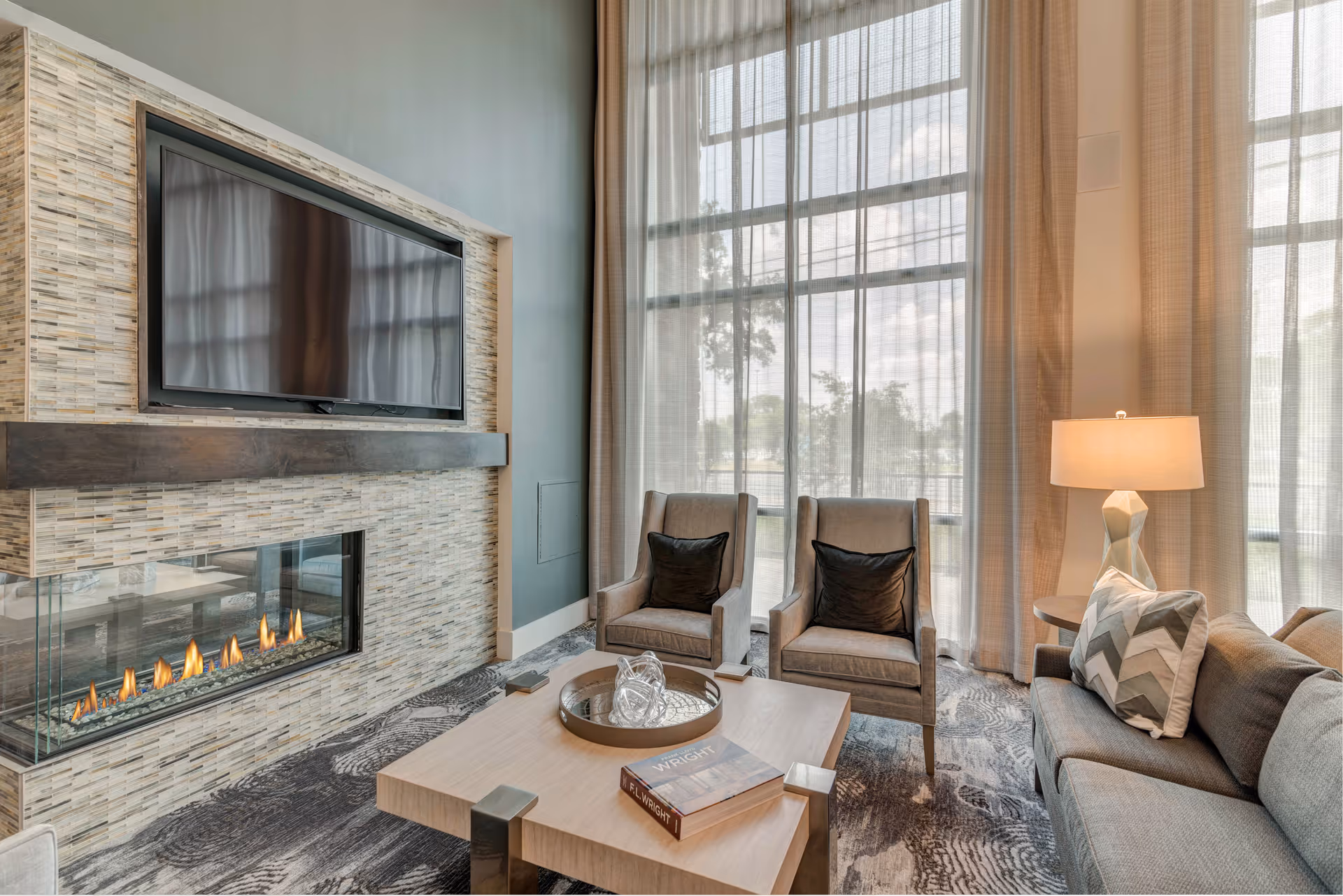 A modern living room with a mounted flat-screen TV above a glass-enclosed fireplace. The room features two gray armchairs with black pillows, a gray sofa with a patterned pillow, a wooden coffee table with a book and decorative glass object, and large windows with sheer curtains allowing natural light to fill the space.