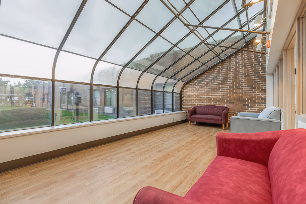 A sunroom with a curved glass ceiling and large windows overlooking a grassy outdoor area. The room has light wood flooring and three sofas in red, purple, and gray colors arranged along the walls. One wall is made of exposed brick.