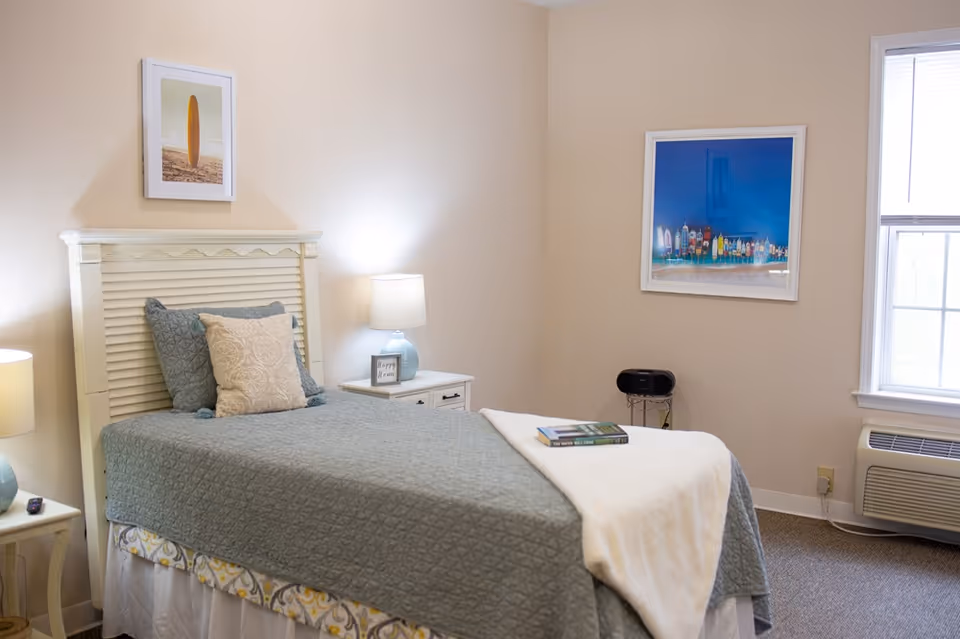 A neatly made single bed with a gray quilt and a white blanket folded at the foot. There are two pillows on the bed, one gray and one beige. The bed has a white headboard. On either side of the bed are small white nightstands with blue lamps. One nightstand has a small framed sign that says 'Happy Home.' The walls are beige with two framed pictures hanging, one of a surfboard on a beach and another colorful artwork. A window with blinds and an air conditioning unit is visible on the right side of the room.