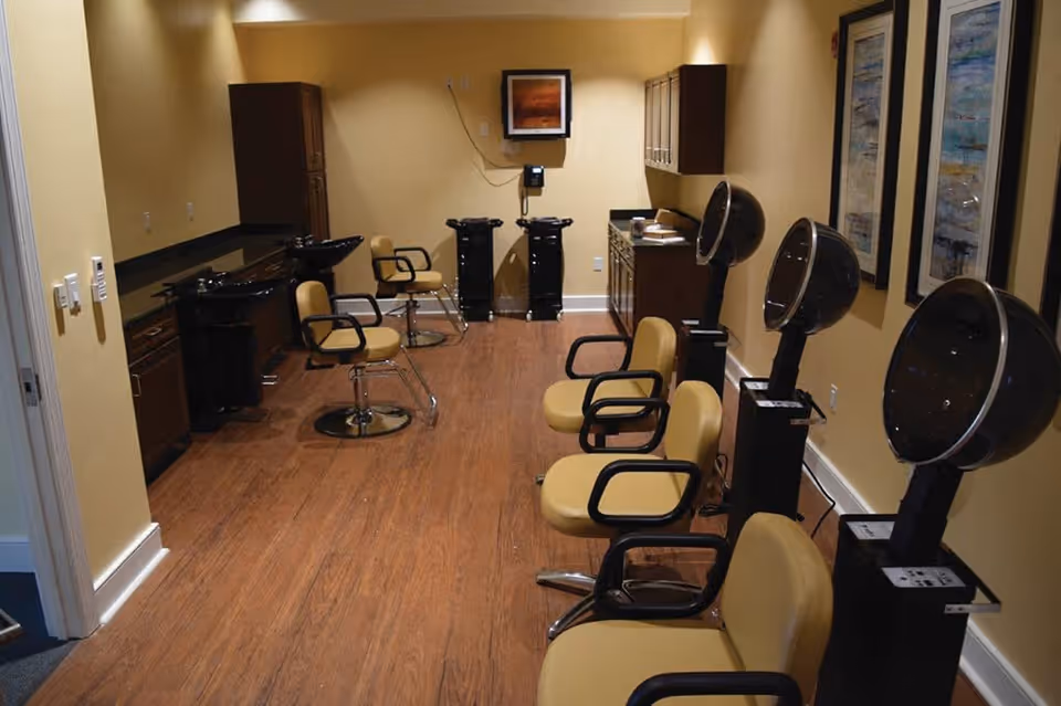 Interior view of a salon area with multiple yellow salon chairs and black hair drying stations along the right wall. On the left side, there are black sinks and additional chairs for hair washing. The room has wooden flooring, beige walls, and framed artwork on the right wall.