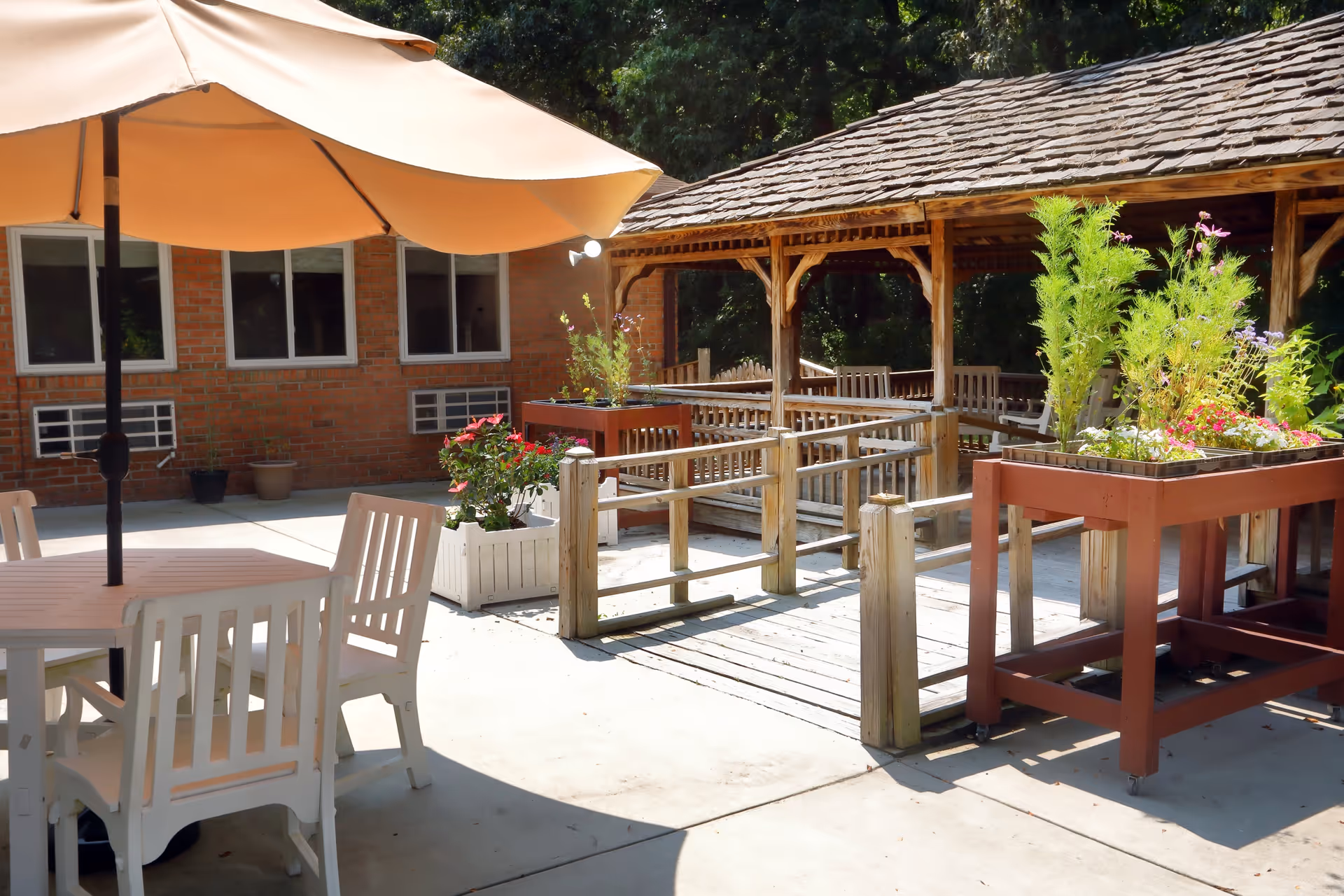 Outdoor patio area with a round table and white chairs under a large beige umbrella, wooden planters with green plants and flowers, a wooden gazebo with benches, and a brick building with windows in the background.