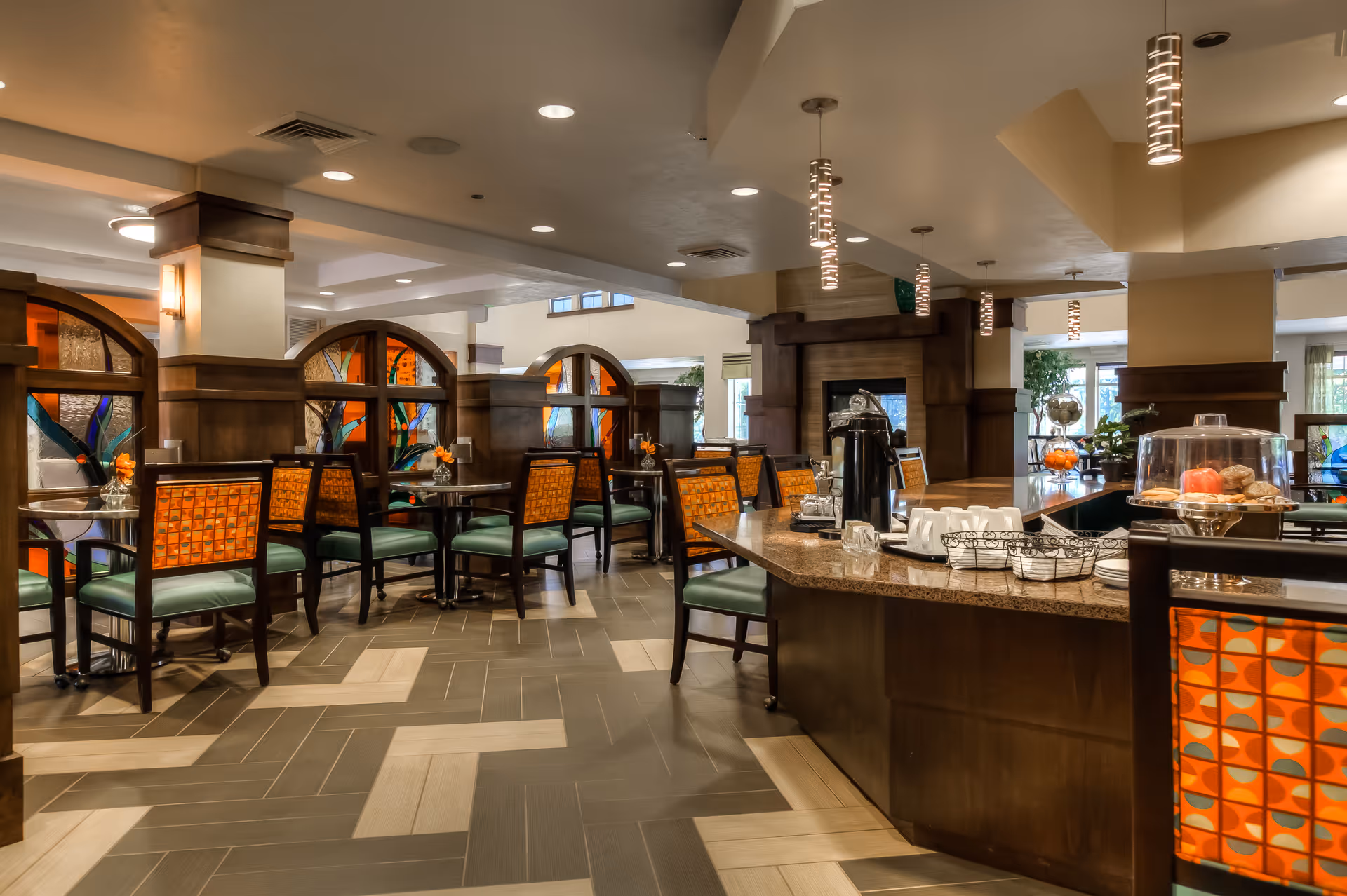 Dining room with tables, patterned chairs and a coffee service counter in a senior living facility.