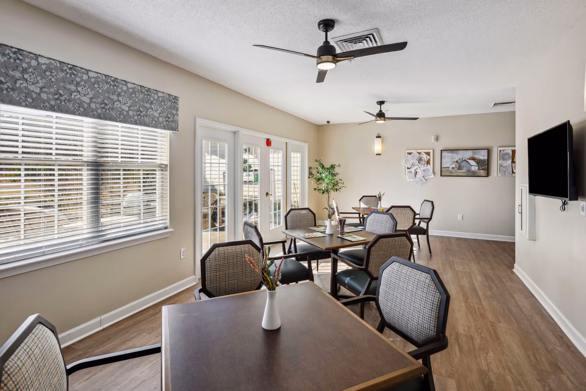 Bright communal dining room with multiple tables and chairs, large windows, ceiling fans, and a wall-mounted TV.