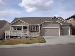 Single-story residential building with a gray roof, beige siding, and stone accents. The house features a front porch with a white railing, a small tree in the front yard, and a concrete driveway leading to a two-car garage.