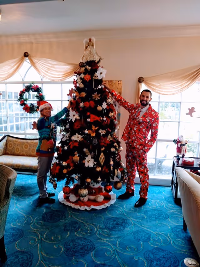 Two people standing on either side of a decorated Christmas tree in a living room. One person is wearing a Santa hat and a festive sweater, while the other is dressed in a red Christmas-themed suit. The room has large windows with sheer curtains, a blue patterned carpet, and couches.