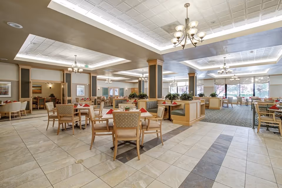 A spacious and well-lit dining room with multiple tables and chairs arranged neatly. Each table has a red folded napkin and a small centerpiece. The room features large windows allowing natural light to enter, decorative ceiling lights, and a tiled floor with a patterned design. There are also some green plants placed on dividers between seating areas.