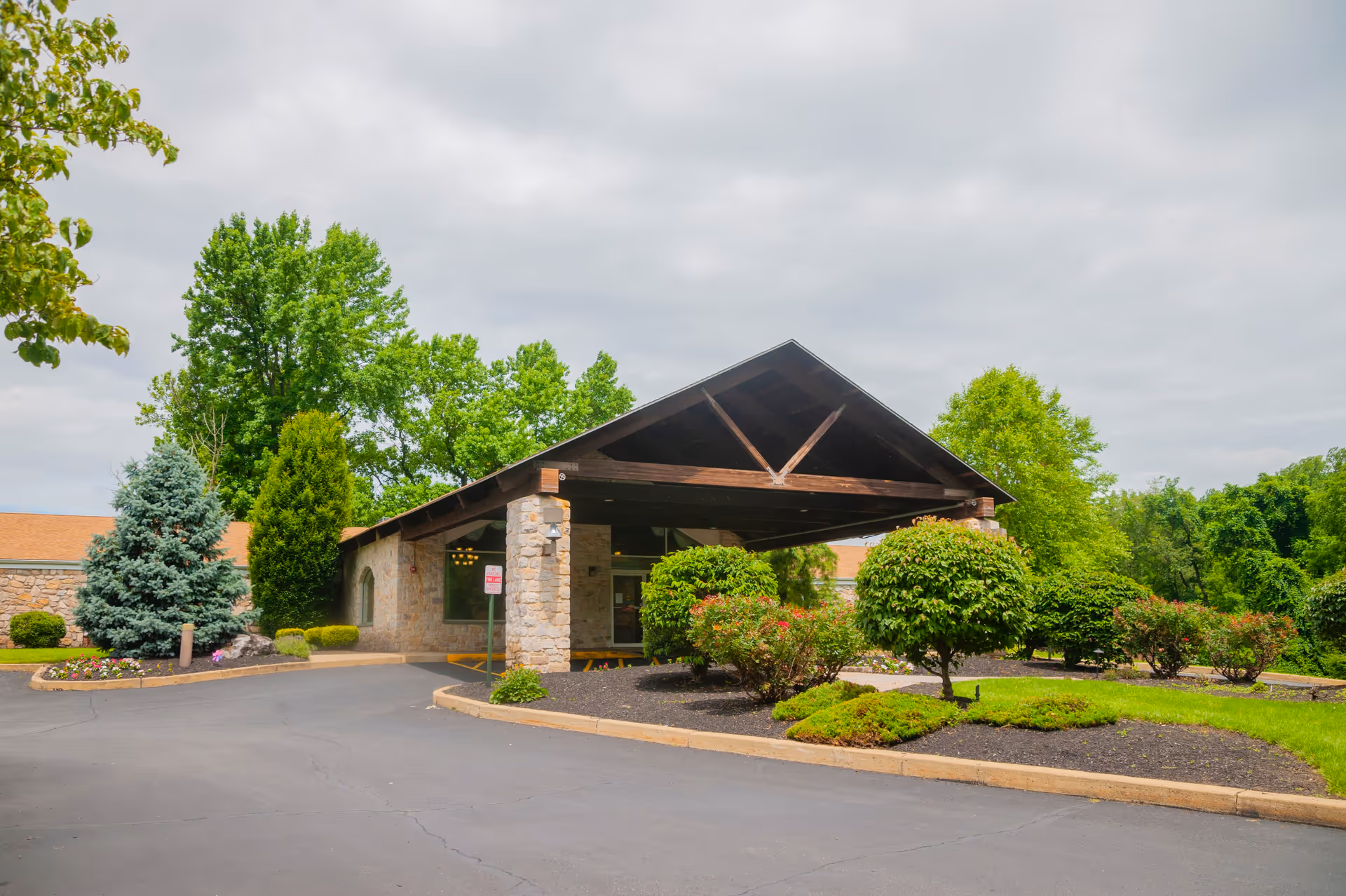 Front entrance of Symphony Manor of Feasterville showing a covered porte-cochere, stone facade, and landscaped shrubs and trees.