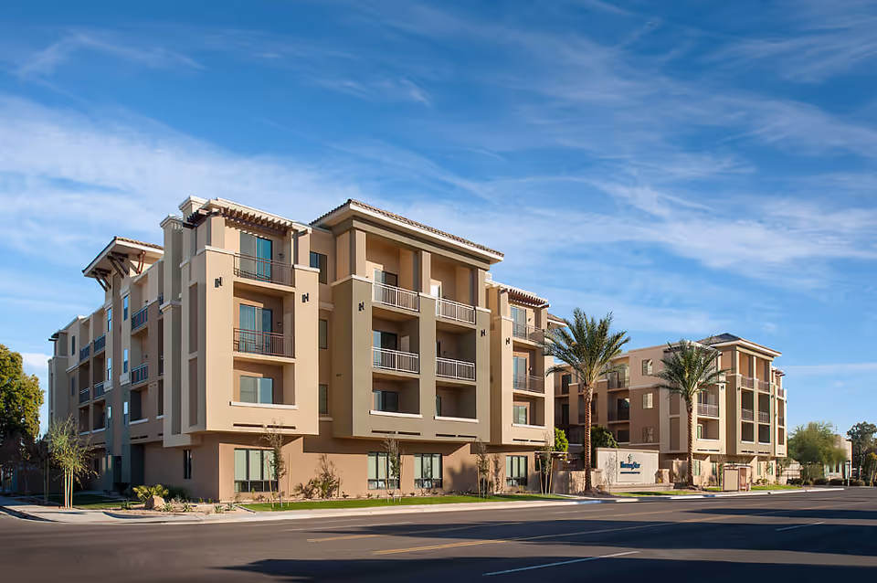 Exterior view of a modern multi-story senior living facility building with balconies, palm trees, and a clear blue sky.