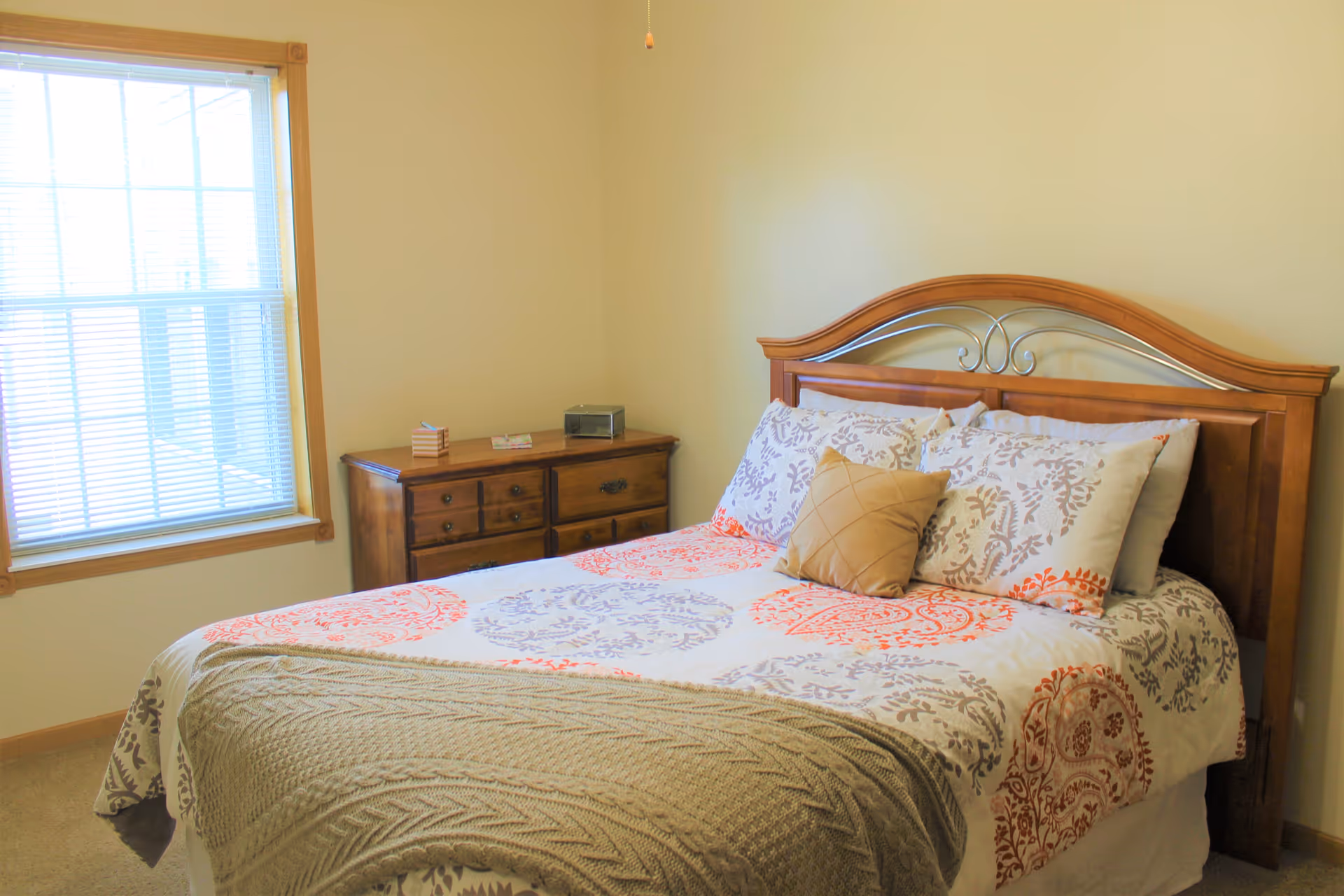 Bright bedroom with a wooden headboard, patterned bedding, a dresser and a window with blinds.