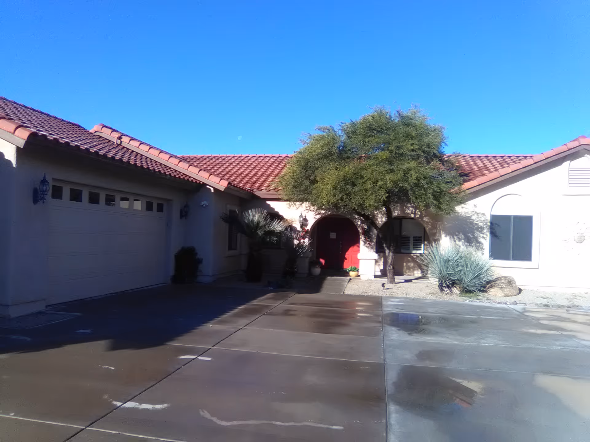 Front exterior of a single-story stucco building with a red tile roof, a red arched entry door, a garage, a tree, and a wet concrete driveway.
