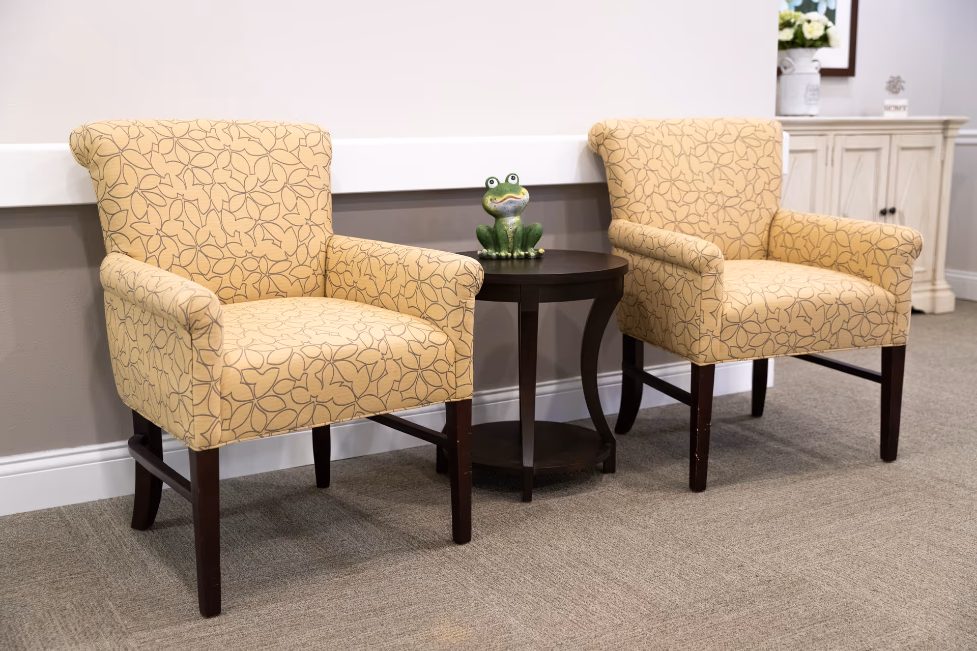 Two beige armchairs with a leaf pattern are positioned on either side of a small round dark wooden table. A small green frog figurine sits on the table. In the background, there is a white cabinet with a flower arrangement on top.