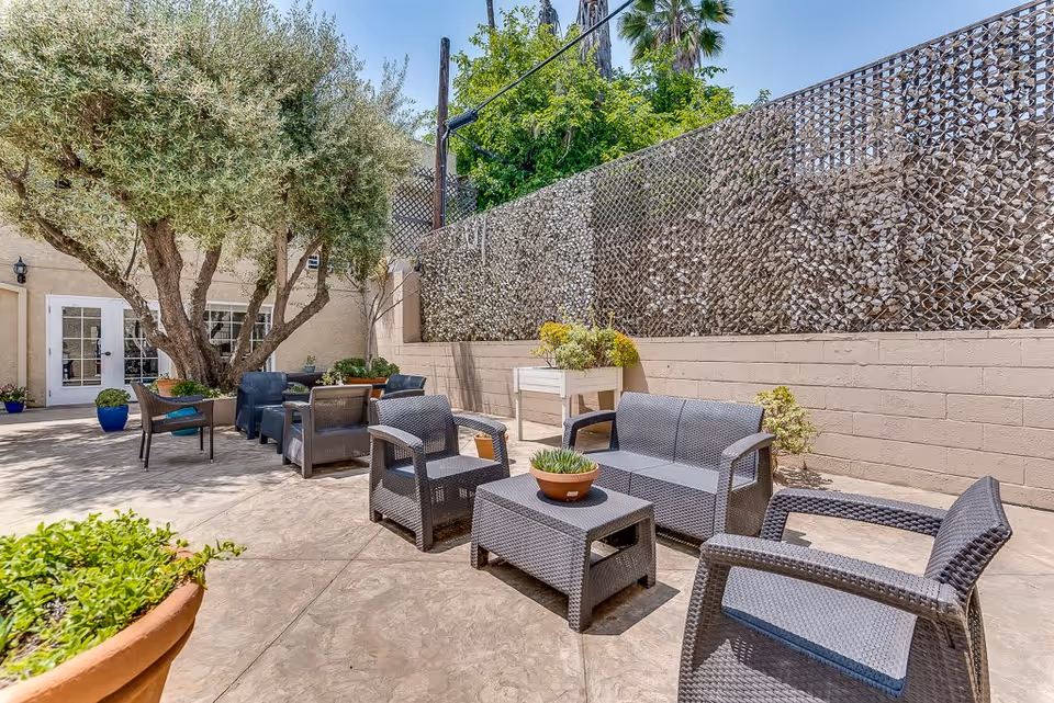Outdoor patio area at Astoria Park Senior Living with several dark wicker chairs and a loveseat arranged around a small table with a potted plant. There are additional seating areas with chairs and tables near a large tree and potted plants along the perimeter. The space is enclosed by a beige wall and a fence covered with foliage.