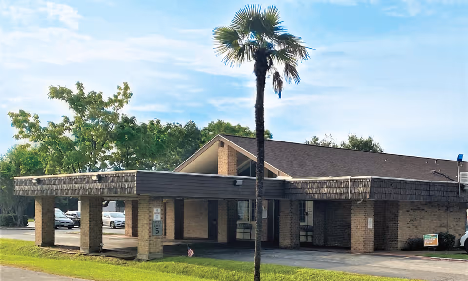 Front exterior of a low brick senior living facility with a porte-cochere and a tall palm tree in front.