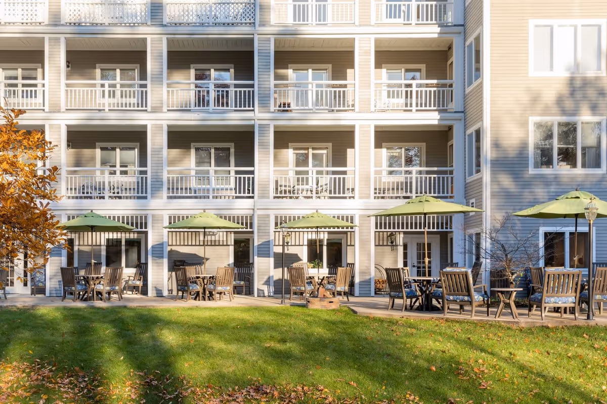 Outdoor patio area of a senior living facility with multiple wooden tables and chairs under green umbrellas, situated in front of a multi-story building with balconies. The area is surrounded by a well-maintained lawn and some autumn-colored trees.