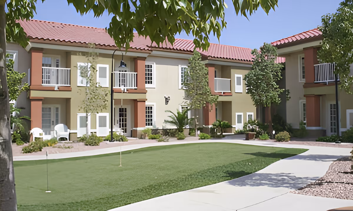 Outdoor courtyard area of a senior living facility with a putting green, surrounded by two-story buildings with balconies and red-tiled roofs, trees, and paved walkways under a clear blue sky.