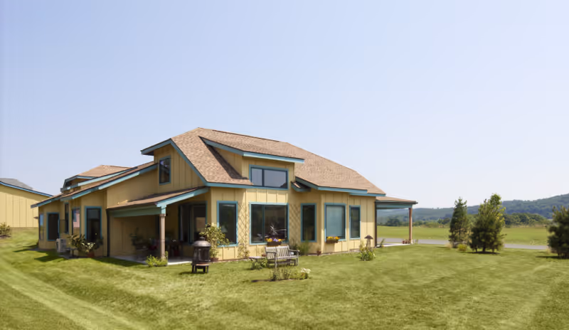 A single-story yellow building with a brown shingled roof situated on a well-maintained grassy lawn. The building has multiple windows and a small covered porch area with some outdoor furniture and plants. The background shows a clear sky and distant trees and hills.