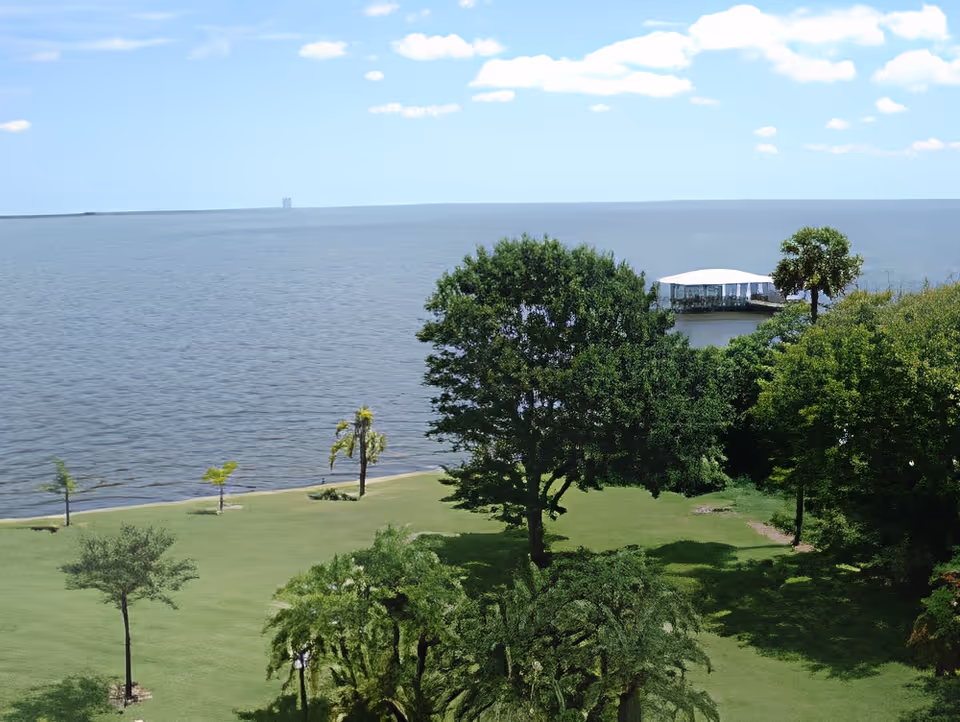 Grassy waterfront lawn with scattered trees and a covered dock on a calm body of water under a blue sky.