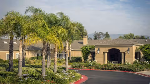 Senior living facility exterior with palm trees, a curved driveway, and an arched entrance.