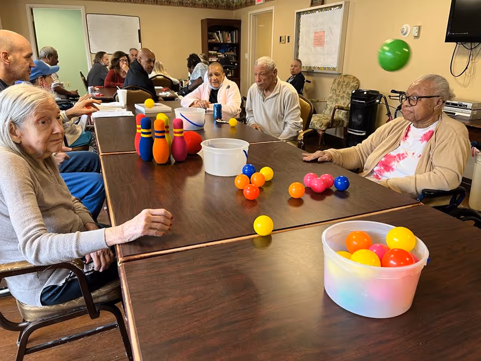 A group of elderly people sitting around a large table in a common room, engaging in a game with colorful plastic balls and bowling pins. The room has beige walls, a bulletin board, and a television mounted on the wall.