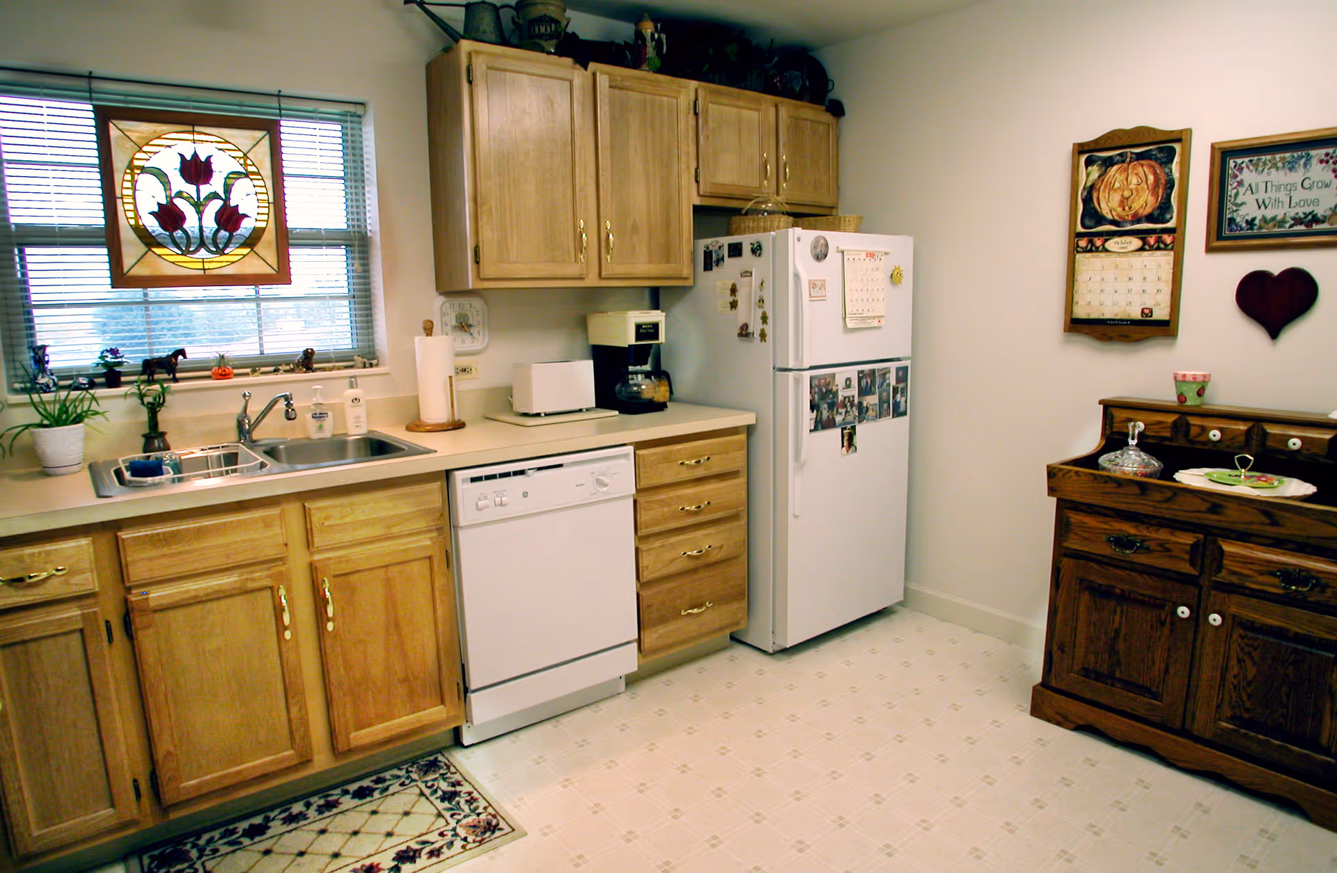 Kitchen with light wood cabinets, a sink under a stained-glass window, white dishwasher and refrigerator, and a wooden sideboard on the right.