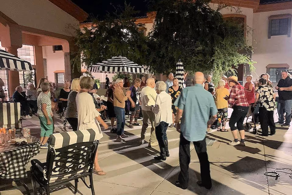 A group of elderly people socializing and dancing outdoors at night in a courtyard area with striped umbrellas and chairs. The setting appears to be a senior living facility with a building visible in the background.