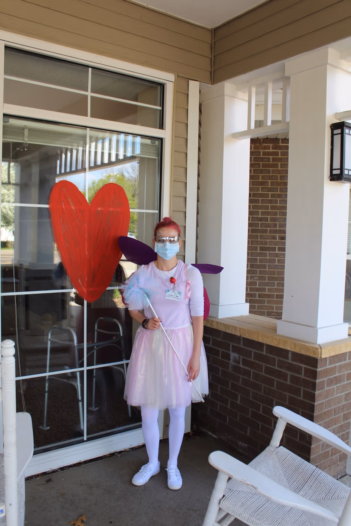 A person dressed in a pink fairy costume with purple wings, white tights, and white shoes stands on a porch outside a building. They are wearing a face mask and protective goggles, holding a wand with a fluffy blue tip. Behind them is a large window with a red heart painted on it. The porch has white rocking chairs and brick walls.