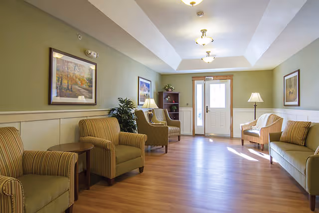 A well-lit living room area with wooden flooring, light green walls, and white wainscoting. The room contains several cushioned armchairs and a sofa arranged along the walls, with side tables and lamps. There are framed paintings on the walls and a potted plant near the chairs. A door with glass panels is visible at the far end of the room.