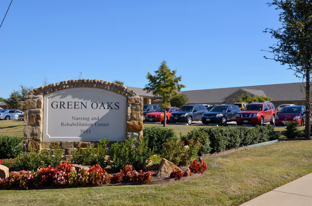 Stone sign for Green Oaks Nursing and Rehabilitation Center with the address 3033, surrounded by landscaping with flowers and bushes, with a parking lot and building in the background under a clear blue sky.