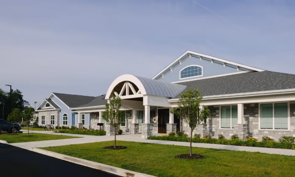 Exterior view of a single-story senior living facility building with a gray shingled roof, light blue siding, and stone accents. The entrance features a white arched canopy supported by columns. There are small trees and landscaped grass areas in front of the building under a clear sky.