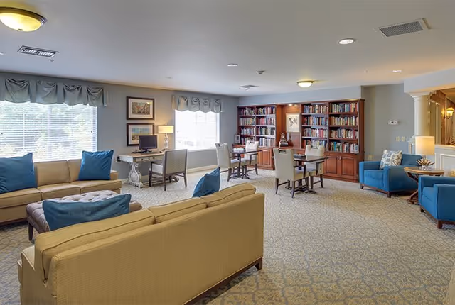 A spacious and well-lit common area with beige sofas adorned with blue cushions, a large window with valances, a desk with a computer, several chairs around tables, and a wooden bookshelf filled with books against the far wall.