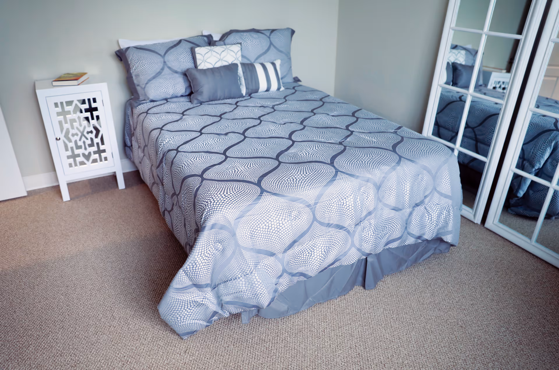 A neatly made bed with a gray and white patterned bedspread and multiple pillows in a small bedroom. Next to the bed is a white nightstand with a book on top. There is a large mirror with a white frame reflecting part of the bed and nightstand. The room has beige carpet and light-colored walls.