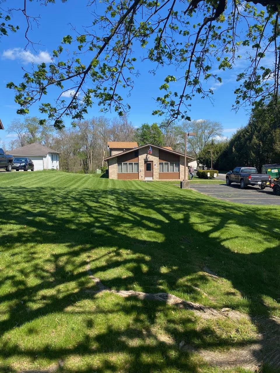A single-story building with a brown roof and beige walls, surrounded by a well-maintained green lawn. There are several vehicles parked on a paved area to the right, and tree branches with green leaves frame the top of the image. The sky is clear and blue with a few clouds.