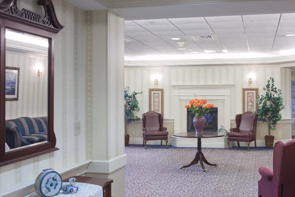 A cozy living room area in Partridge House featuring two brown leather armchairs positioned on either side of a white fireplace. A round wooden table with a vase of orange flowers is centered in front of the fireplace. The room has patterned carpet flooring, wallpapered walls with framed artwork, and potted plants on either side of the fireplace. A large mirror on the left wall reflects part of a blue patterned sofa.