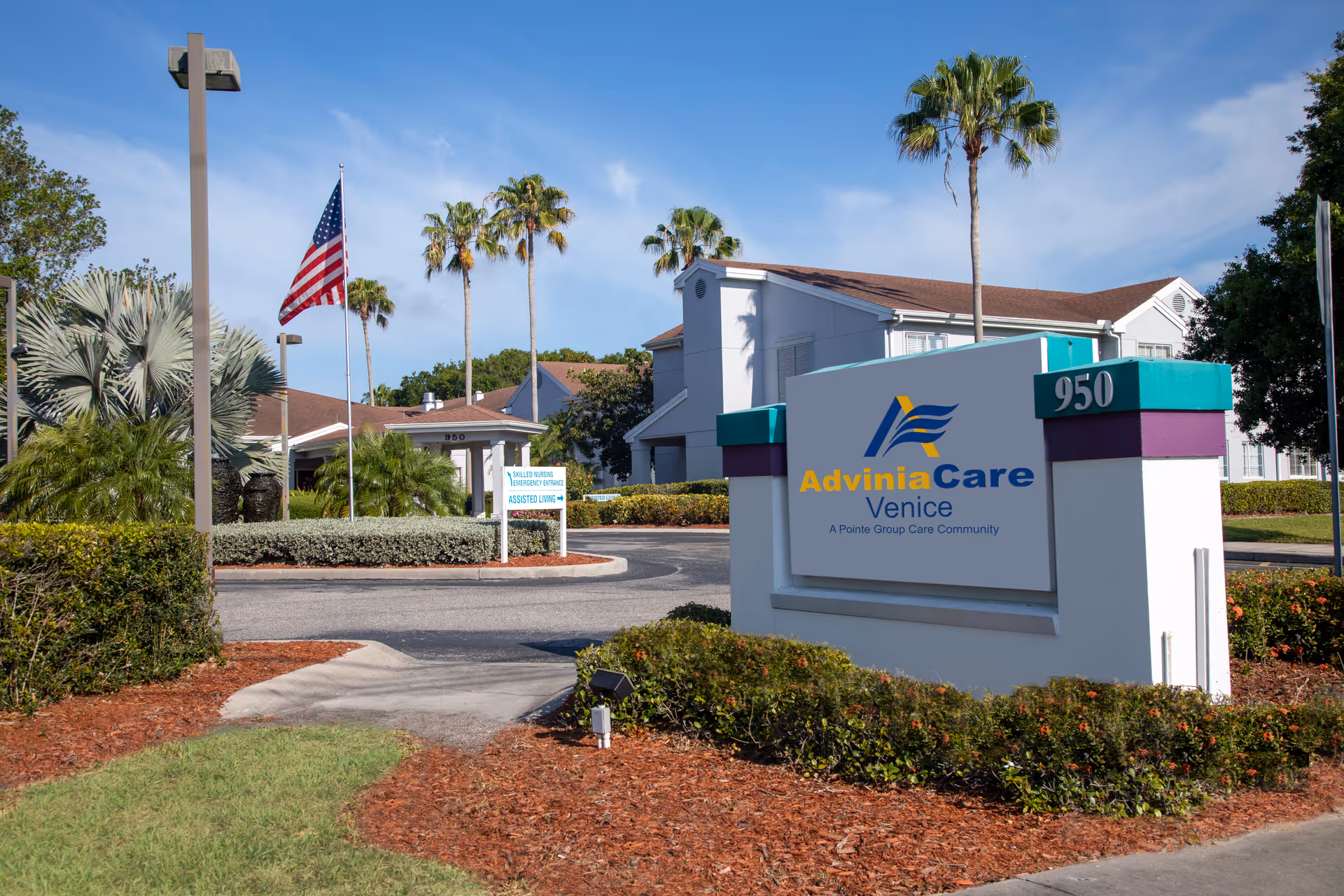 Exterior view of AdviniaCare Venice senior living facility with a large sign displaying the facility name and address number 950. The building is surrounded by palm trees, bushes, and a clear blue sky. An American flag is flying on a flagpole near the entrance.