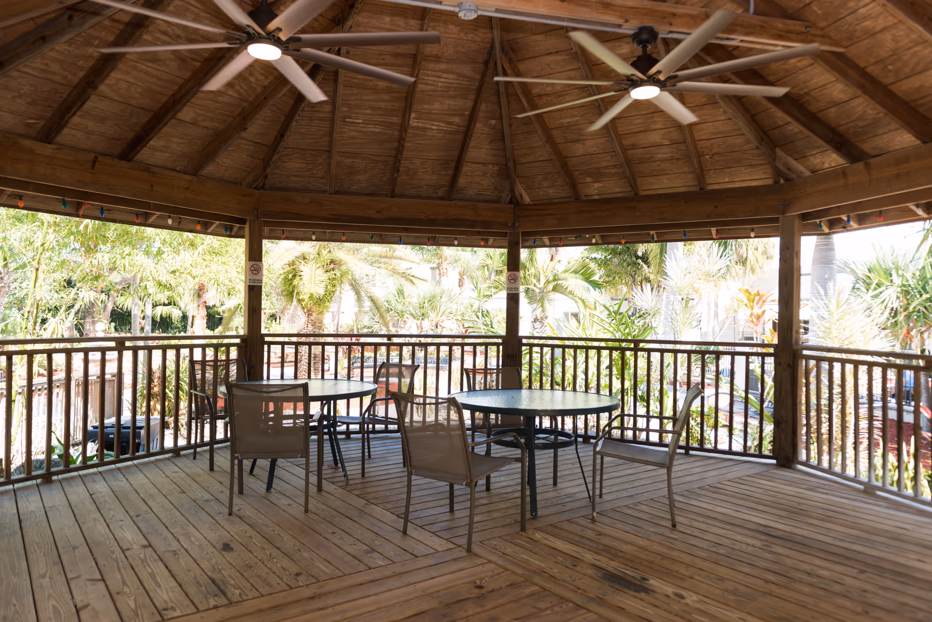 A wooden gazebo with a high ceiling and two ceiling fans, containing two round tables each surrounded by four chairs. The gazebo is open on all sides with wooden railings, and lush greenery and palm trees are visible outside.