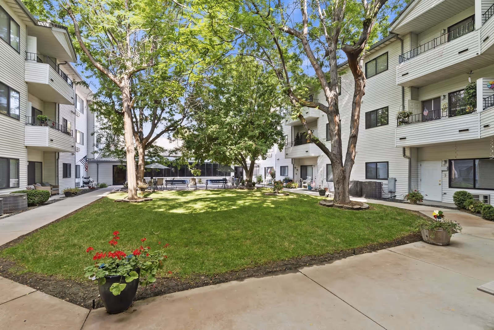 Outdoor courtyard area of a senior living facility with green grass, several large trees, potted plants with red flowers, and surrounding multi-story white buildings with balconies and windows.