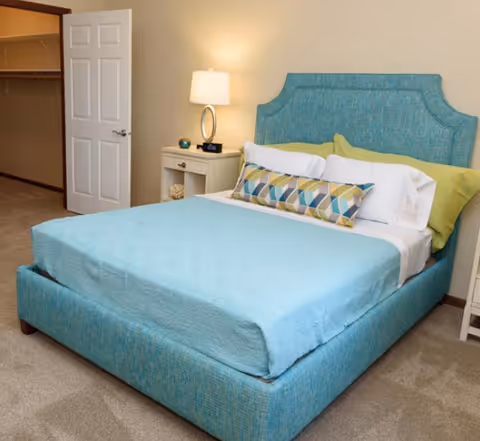 A bedroom with a blue upholstered bed featuring a matching headboard and bed frame, neatly made with a light blue bedspread, white and green pillows, and a decorative geometric-patterned bolster pillow. A nightstand with a lamp, alarm clock, and decorative items is beside the bed. An open door reveals a walk-in closet with shelving and hanging rods.