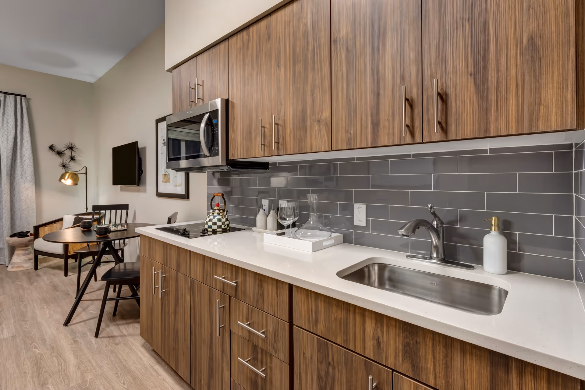 Modern studio kitchen with wood cabinets, gray subway tile backsplash, white countertop with sink and microwave, and a small dining table in the background.