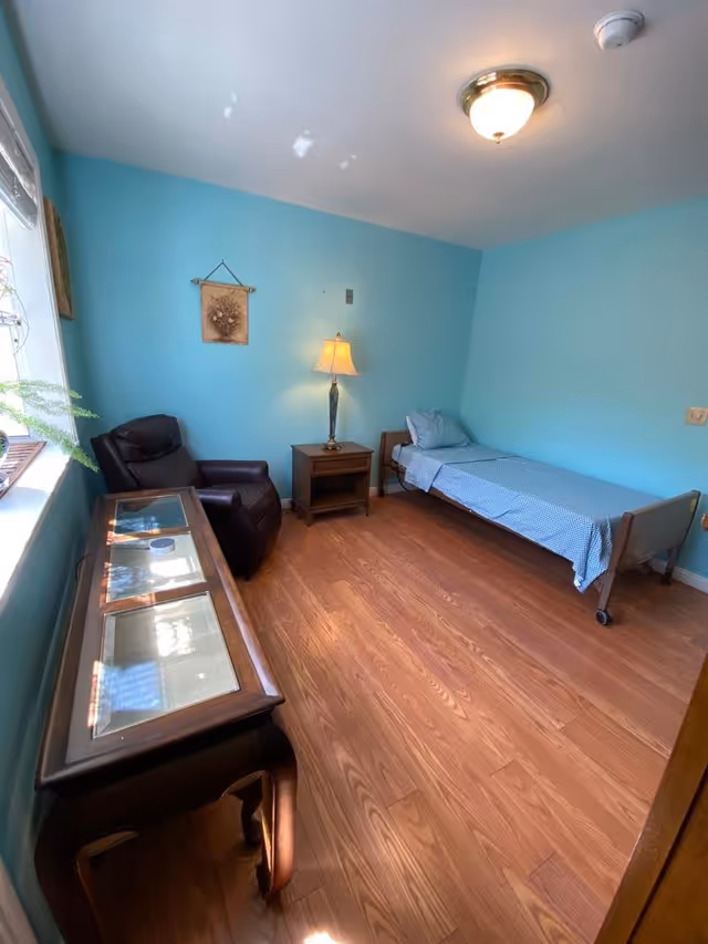 A small bedroom with light blue walls and wooden flooring. The room contains a single bed with blue checkered bedding, a wooden nightstand with a lamp, a dark brown recliner chair, and a wooden table with glass panels on top. There is a window on the left side letting in natural light and a small wall decoration above the nightstand.