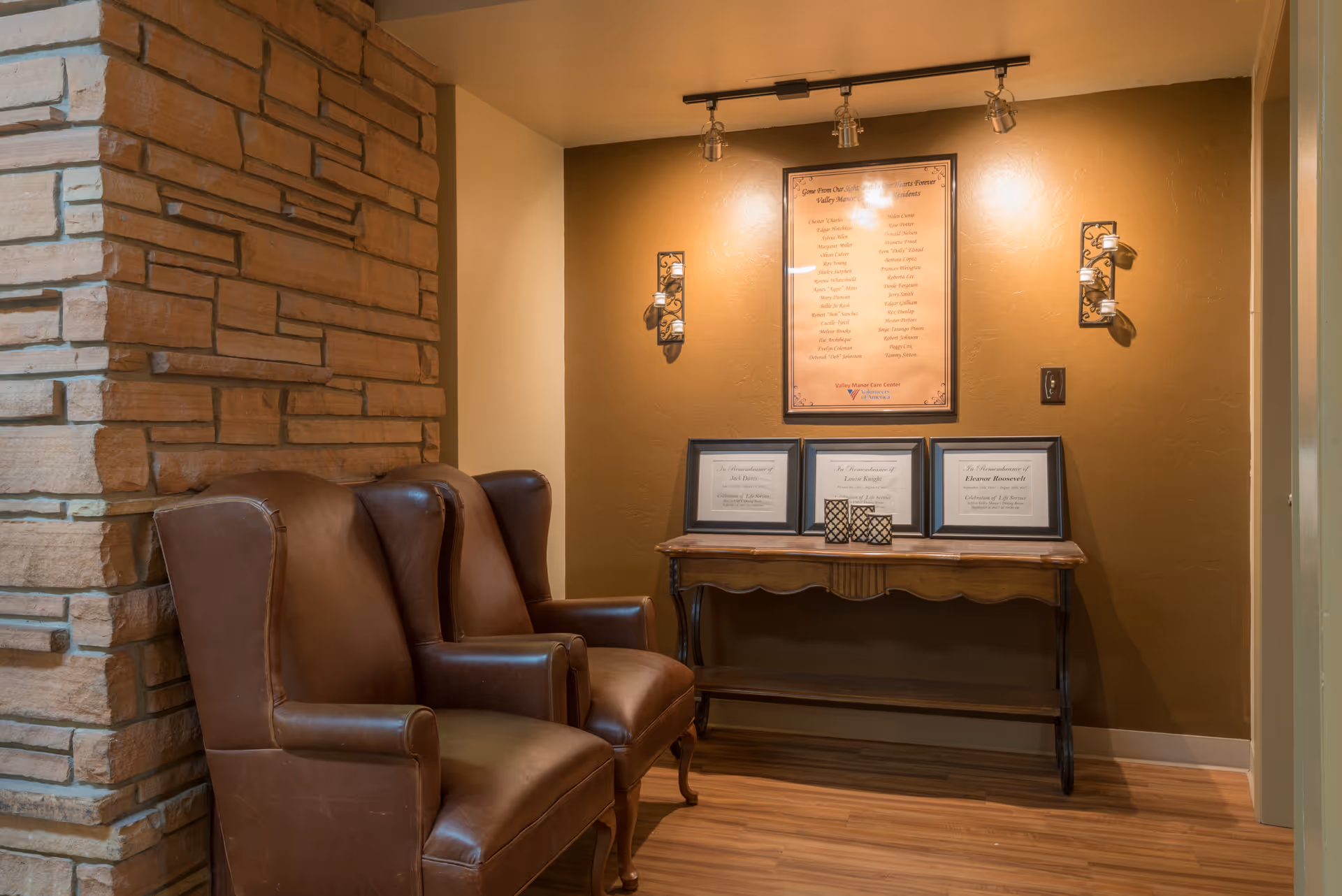 A cozy corner with two brown leather armchairs next to a stone wall. On the opposite wall, there is a wooden console table holding three framed certificates and two decorative candle holders. Above the table, a large framed plaque is mounted on a brown wall, illuminated by three ceiling spotlights and two wall sconces.
